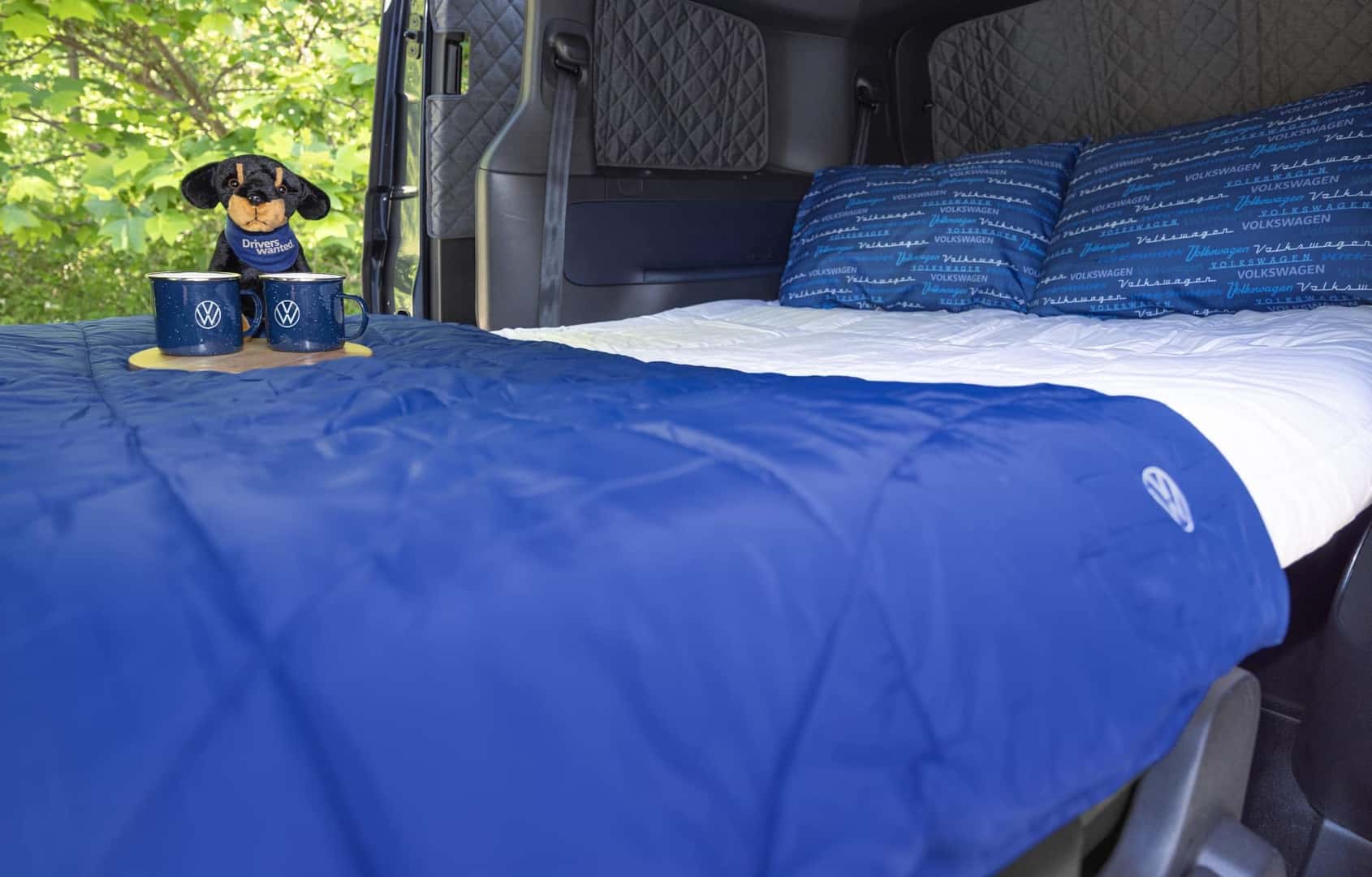 A Cushioned Bed With Blue And White Bedding Inside The Spacious Volkswagen Atlas, With An Open Door. On The Bed, There Is A Cute Stuffed Dog Wearing A 'Drivers Wanted" Bandana and Two Volkswagen Mugs