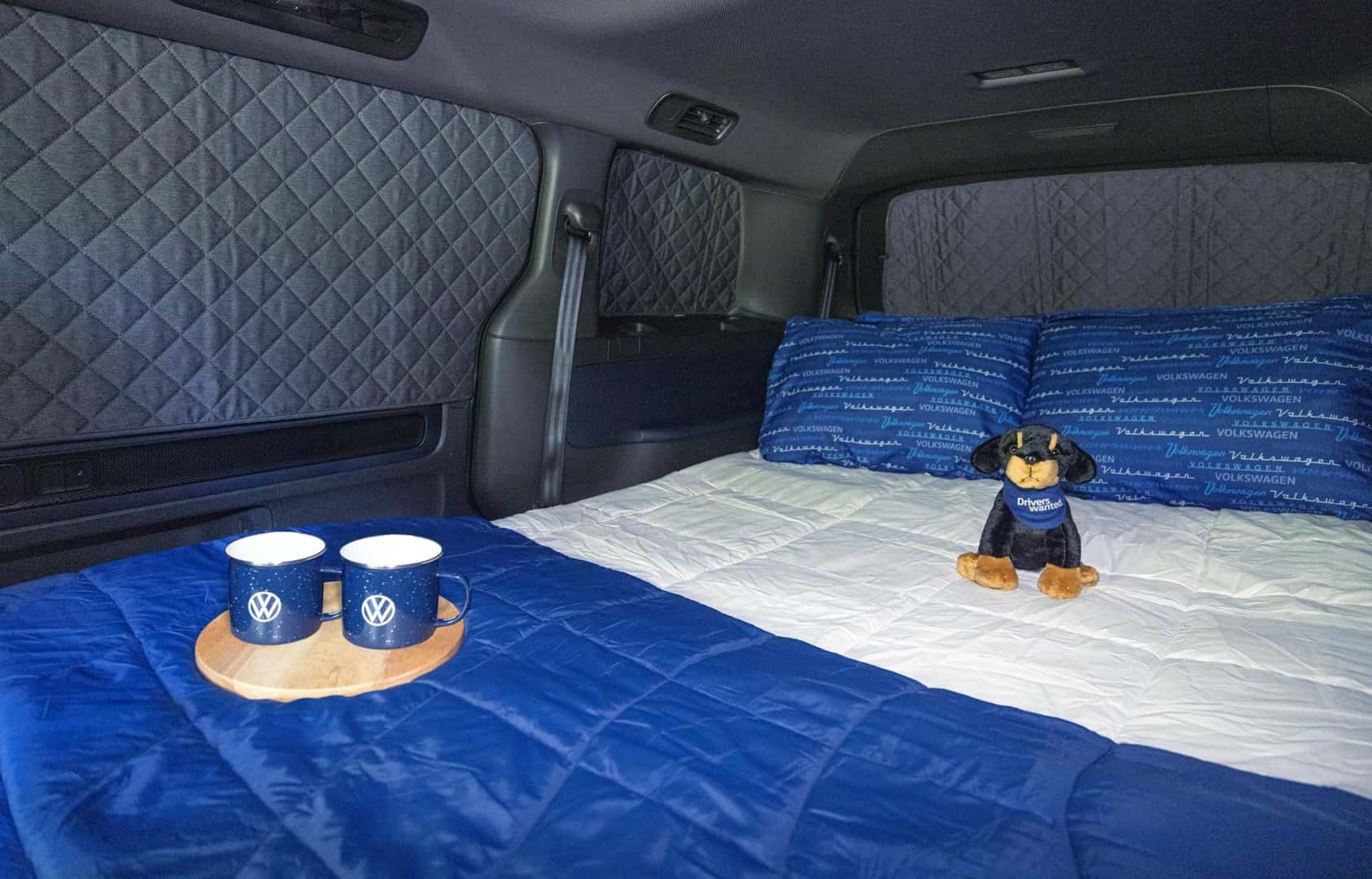 A Cushioned Bed Inside The Volkswagen Atlas, Which Has Covered Windows, Has A Blue And White Bedding, A Cute Stuffed Animal Wearing A 'Drivers Wanted' Bandana, and A Circular Wooden Board Holding Two Volkswagen Mugs