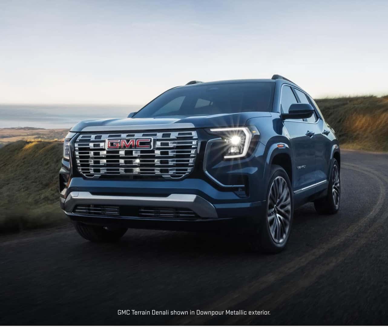 Front three-quarter view of a 2027 GMC Terrain Denali in Downpour Metallic driving along a coastal cliffside road at sunset.