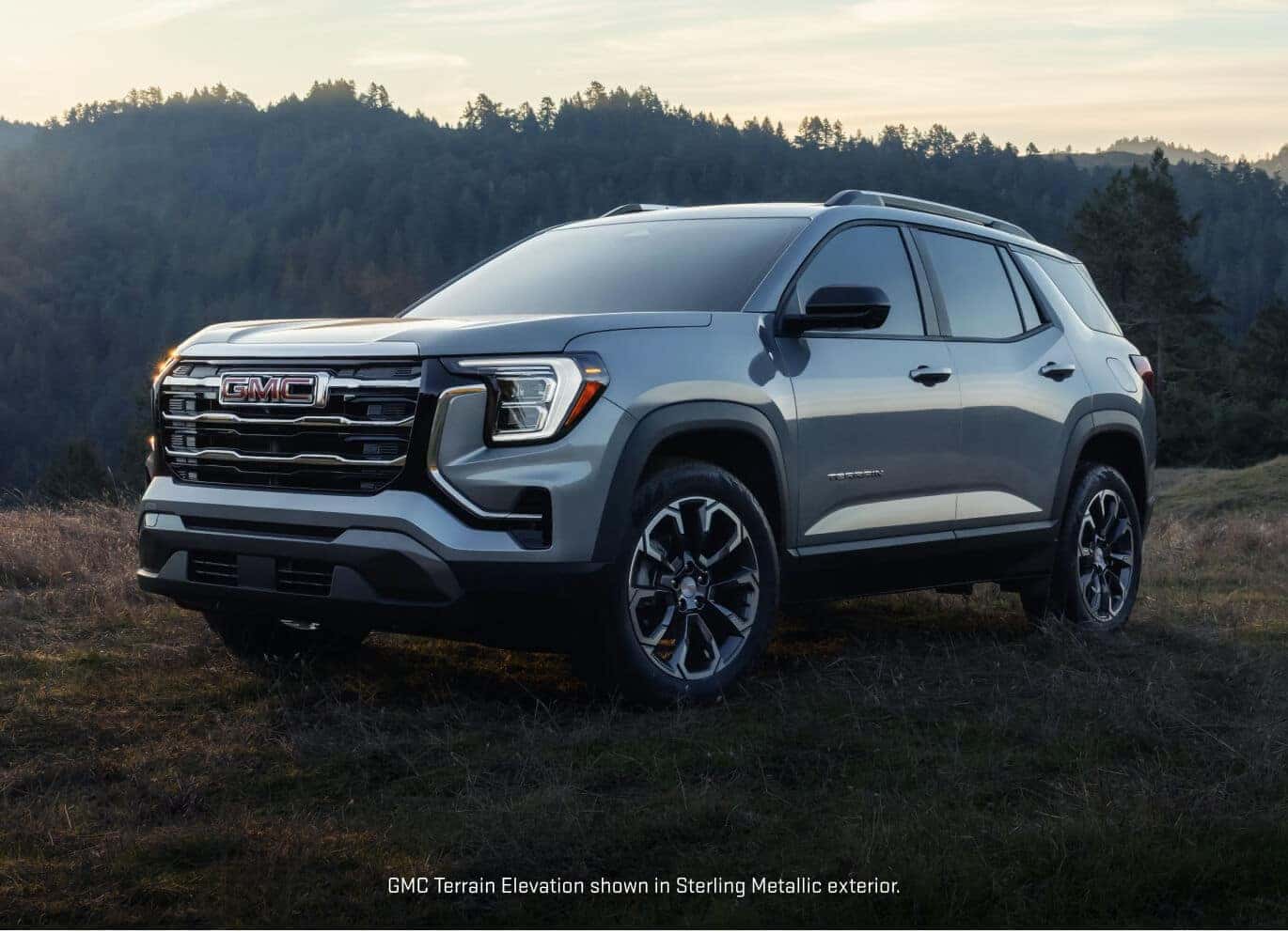 Front three-quarter view of a 2027 GMC Terrain Elevation in Sterling Metallic parked on a grassy ridge against a forested mountain sunset backdrop.