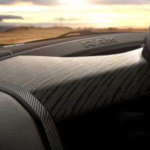 close up of the dashboard of a ram vehicle featuring textured woodgrain panel, RAM badging, and part of the infotainment screen with control buttons.