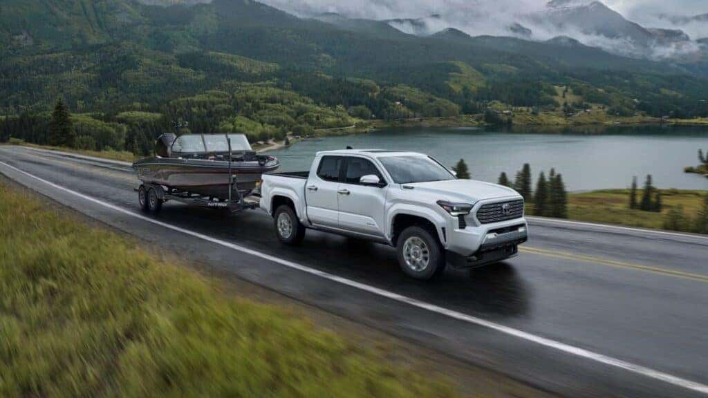 White Toyota Tacoma towing a fishing boat along a scenic lake road surrounded by lush greenery and mountains.