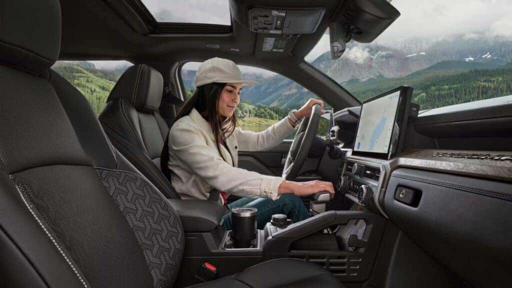 Interior of a Toyota Tacoma with a female driver adjusting controls, showcasing the dashboard and a scenic mountain view outside.