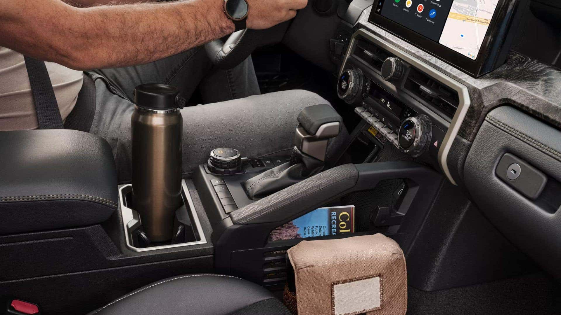 Close-up of the center console in a Toyota Tacoma with a stainless steel travel mug, gear shifter, and a storage slot holding a guidebook.