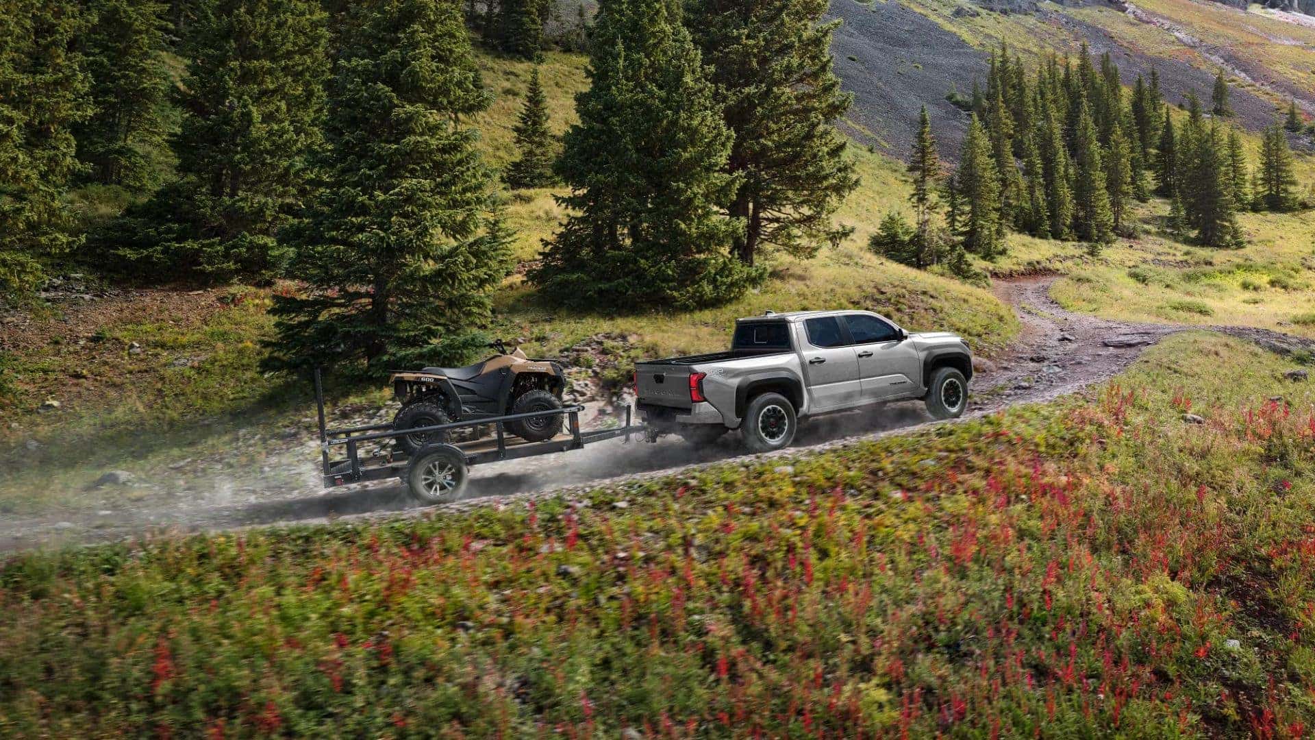 Rear view of a Toyota Tacoma pulling an ATV trailer through a rugged forest trail.
