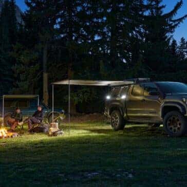Toyota Tacoma parked at a campsite at night, with people gathered around a campfire under the awning.