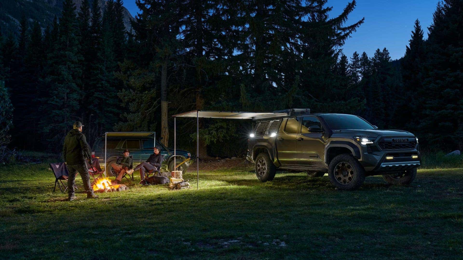 Toyota Tacoma parked at a campsite at night, with people gathered around a campfire under the awning.