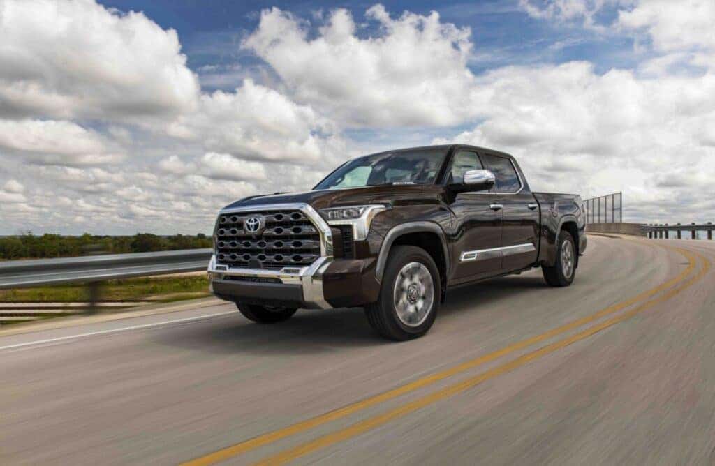 a brown toyota truck driving on a bridge next to a body of water and train tracks