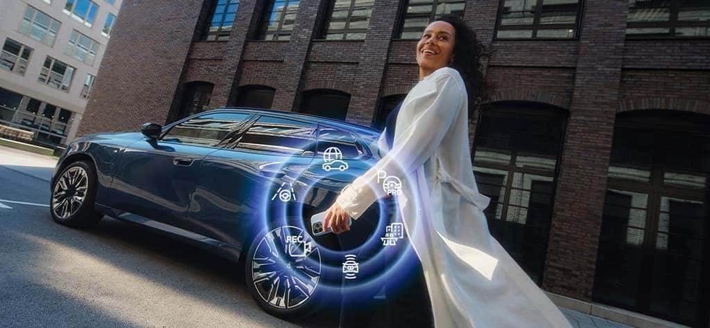 A woman stands in from of a BMW X3 holding a phone bursting with BMW ConnectedDrive icons.