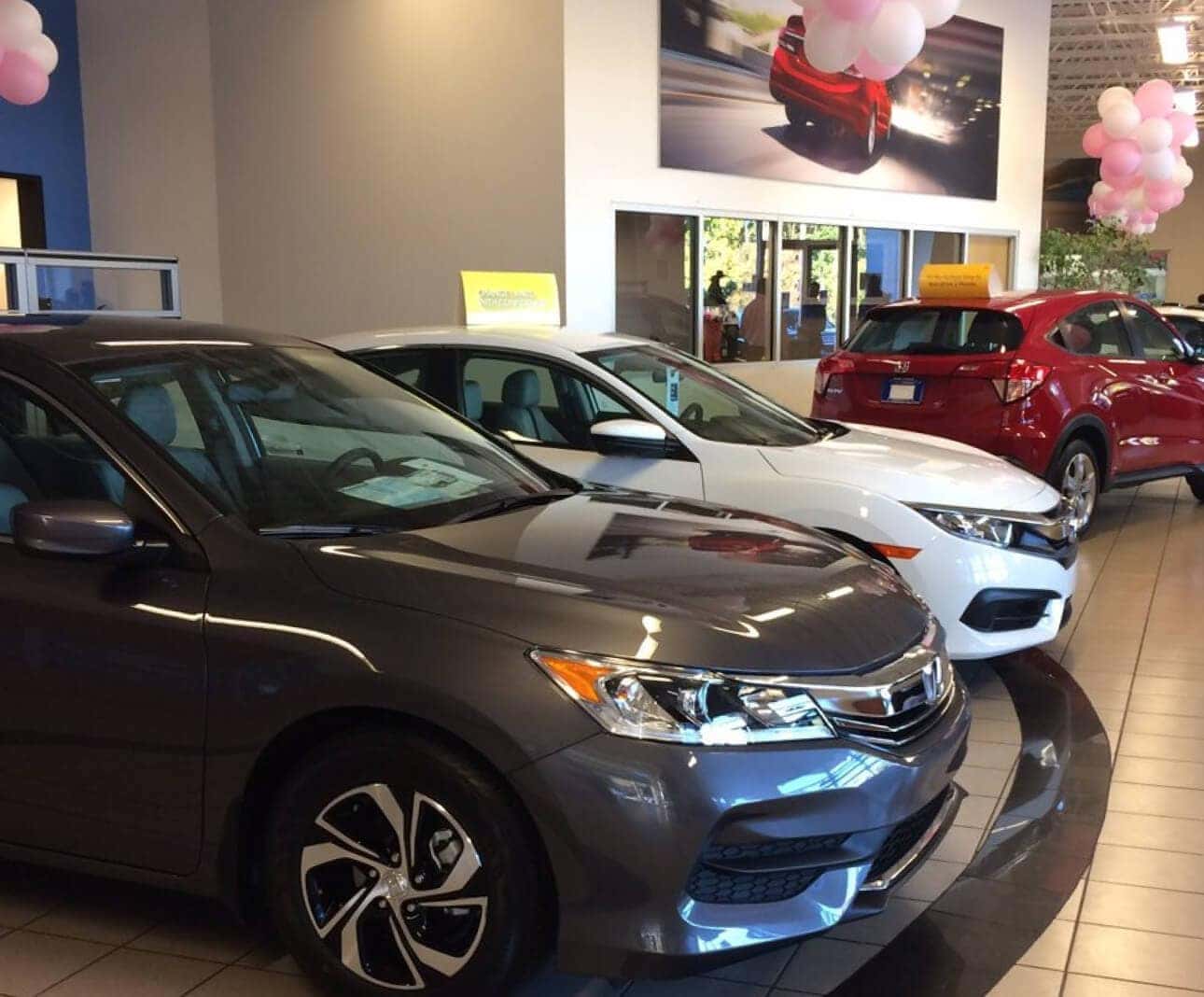 Interior of SONS Honda dealership with a line of used Honda vehicles displayed with balloons.