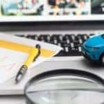 a laptop with a pen and notes atop it, along with bills of money, a magnifying glass, and a toy car